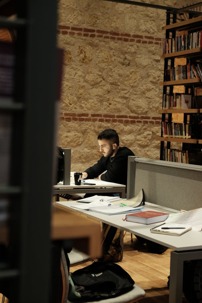 Man seated at a library desk studying amid books and notes. Warm, inviting atmosphere.