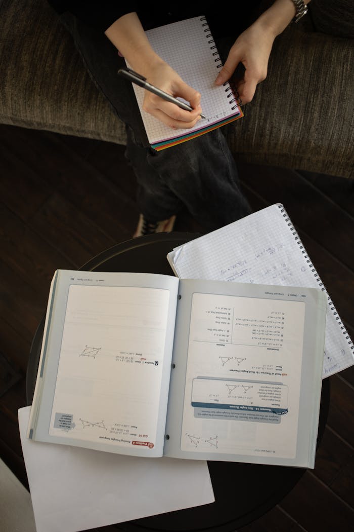 Top view crop faceless person taking notes in planner while sitting on sofa near table with opened notebooks
